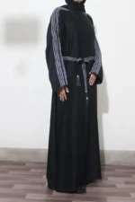 Woman in a black abaya design for women with white striped embellishments and tassel belt, side pose with hands in front.
